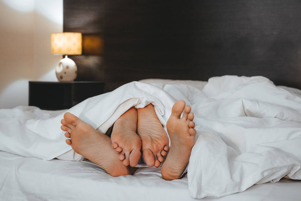 пара ног, выглядывающих из-под белого одеяла на темной кровати молодоженов медового месяца - Фото, изображение