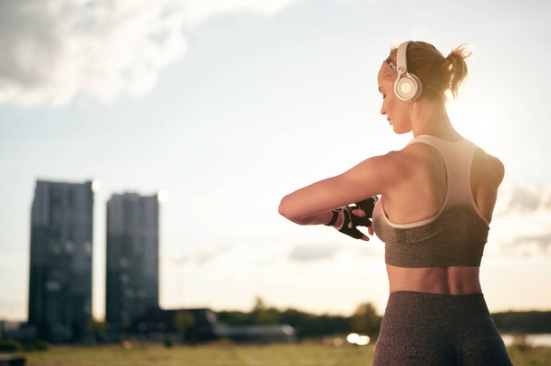 Rückenansicht einer kräftigen, muskulösen Frau in Sportbekleidung und Kopfhörer, die die Smartwatch anpiepst und eine mobile Anwendung nutzt, Hintergrundbeleuchtung, Stadtansicht - Foto, Bild