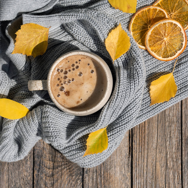 Φθινόπωρο ή Φθινόπωρο πρωινό καφέ. Φθινοπωρινά κίτρινα φύλλα, πατατάκια πορτοκάλι, ζεστή κούπα καφέ στον ατμό και ένα άνετο γκρι πουλόβερ σε ξύλινο φόντο. Κυριακή χαλάρωση, φθινοπωρινή διάθεση και ήρεμη ζωή έννοια. - Φωτογραφία, εικόνα
