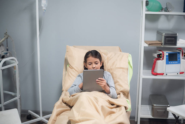 Маленька дівчинка відпочиває в ліжку в лікарняному палаті, вона грає в ігри на своєму цифровому планшеті
 - Фото, зображення