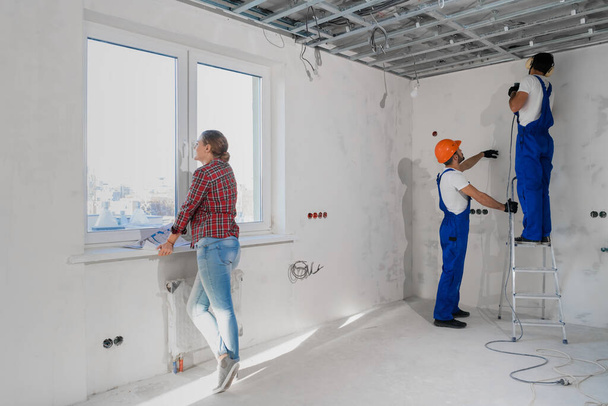 The female client examines the layout of the apartment, general laborers are drilling the wall on a stepladder - Valokuva, kuva