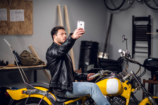 A stylish guy on a yellow cool motorcycle takes a selfie in a garage or car repair shop - Foto, imagen