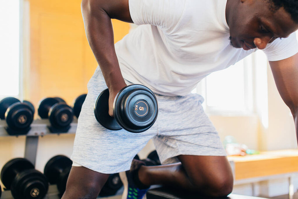 Side view of African American man in sportswear leaning on bench and doing row exercise with dumbbell during training in gym - Fotografie, Obrázek