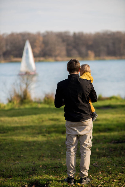 Dad holds his son in a yellow jacket in his arms, they stand with their backs to the camera and look at the lake. Autumn park, Indian summer. - Фото, изображение