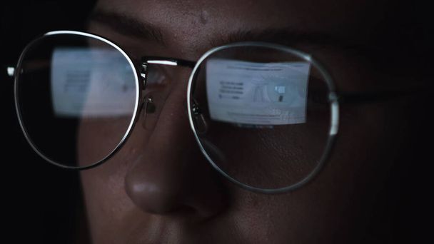 Woman use laptop computer and reading an article at night. Concept. Close up of young female face with big glasses and the reflection of the monitor in lenses isolated on black background. - Photo, Image