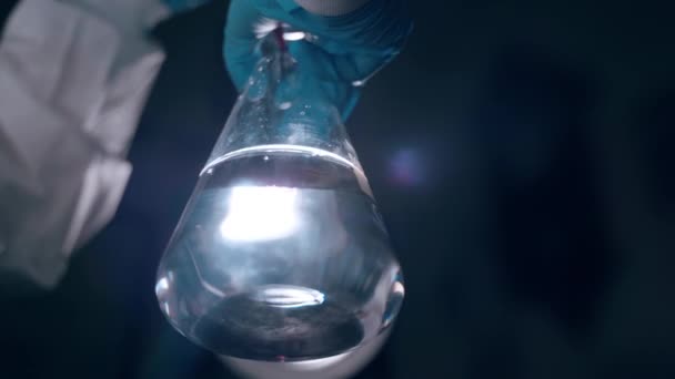Nízkoúhlý záběr skleněné baňky s průhlednou látkou na pozadí kontrastního světla. Lékaři ruku v ochranné modré rukavici kapající červený roztok z odměrné pipety do lékařské baňky pro - Záběry, video