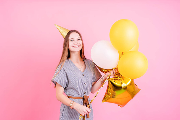 Fiesta de cumpleaños. Mujer joven en un sombrero de cumpleaños sosteniendo globos y caja de regalo grande celebrando fiesta de cumpleaños sobre fondo rosa con espacio para copiar - Foto, imagen