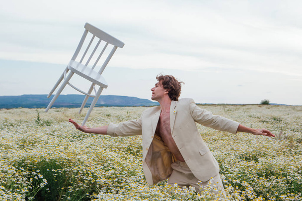 Tall handsome man standing in the middle of camomile flowers field and balancing with white chair on his palm - Φωτογραφία, εικόνα