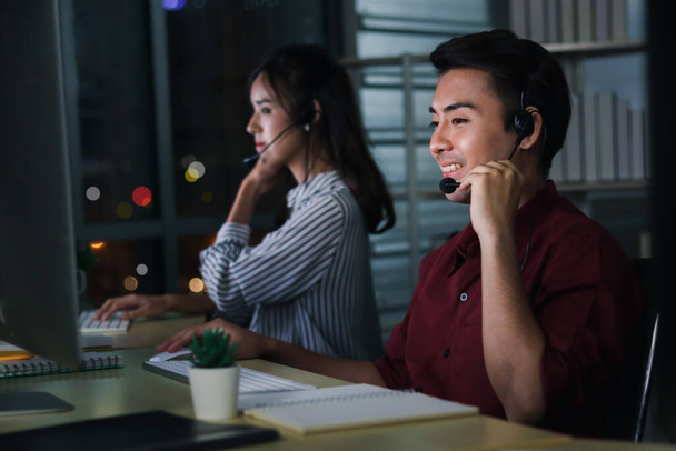 Happy smiling young Asian man with headphones working at call center service desk consultant with her teammates at night, taking with customer on hands-free phone, happy workplace concept  - Photo, Image