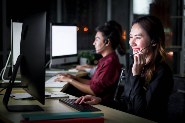 Happy smiling young beautiful Asian woman with headphones working at call center service desk consultant with her teammates at night, ready to take with customer on hands-free phone, happy workplace concept - Фото, изображение