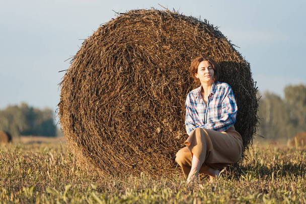 Romantische brunette vrouw in een geruit shirt en broek zittend op een ronde hooiberg in een geweldig veld, op de achtergrond bos. Warme kleuren van de zonsondergang. - Foto, afbeelding