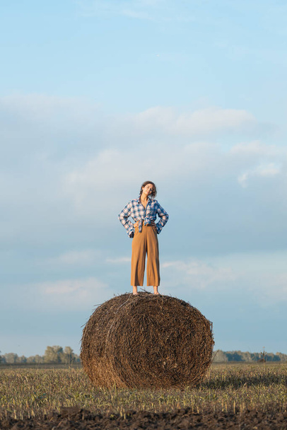 Front view of romantic woman with curly hair in a plaid shirt and trousers smiling and  dancing on a round haystack and relaxing on a meadow on a sunny summer day - Foto, Imagem