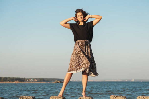 Pozowanie kobiety w naturze. Młoda kobieta w stylowych ubraniach: top i spódnica spacerująca po kamieniach na tle morza. Zdjęcia z wakacji letnich  - Zdjęcie, obraz