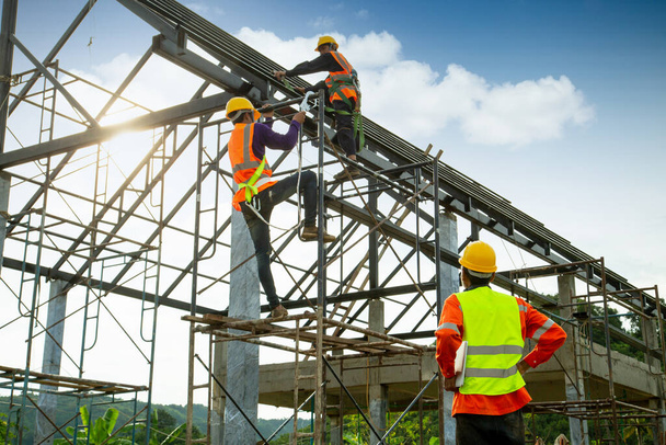 Asiatische Bauarbeiter kontrollieren die Konstruktion von Dachkonstruktionen und Arbeiter tragen Sicherheitshöhen, um das Dach zu installieren. Sturzsicherung für Arbeiter mit Haken für Sicherheitsgurt, Arbeiter wie auf der Baustelle sitzen - Foto, Bild