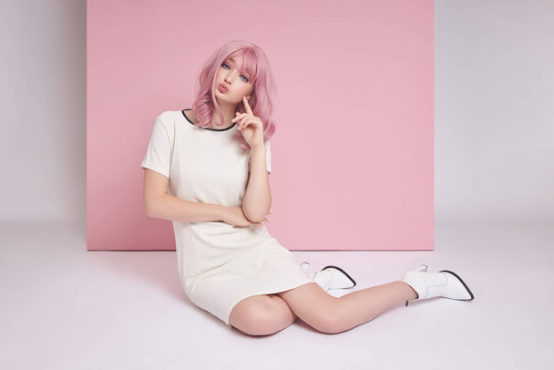 Beautiful girl with long pink hair sitting on the floor, hair coloring. Cute woman on a pink background in a short white dress. Colored hair, perfect hairstyle - Fotó, kép