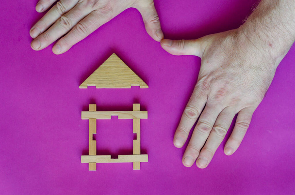 Las manos están protegiendo una pequeña casa de madera de juguete. Las manos de un hombre adulto protegen una casa de madera sobre un fondo púrpura. Seguro de bienes raíces. Protección de la propiedad privada. - Foto, Imagen
