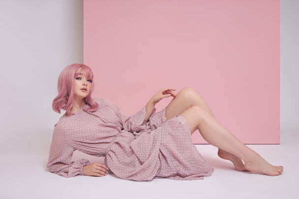 Woman with dyed pink hair in a long dress is sitting on the floor. Portrait of a girl with hair coloring at the pink wall. Perfect hairstyle and hair styling - Foto, afbeelding