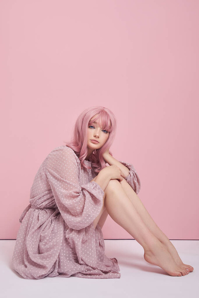 Woman with dyed pink hair in a long dress is sitting on the floor. Portrait of a girl with hair coloring at the pink wall. Perfect hairstyle and hair styling - 写真・画像