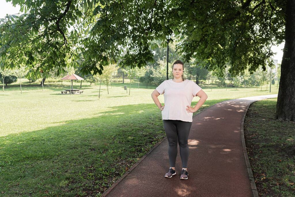 Fat woman standing with arms akimbo on garden path during workout - Photo, Image
