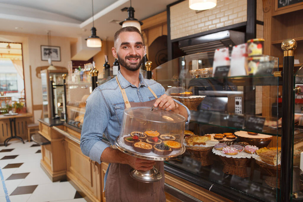 Feliz panadero guapo sosteniendo bandeja con tarteletas deliciosas para la venta en su panadería - Foto, imagen