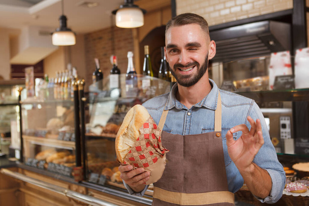 Cheerful male baker smiling, showing ok sign while selling delicious bread - Photo, Image