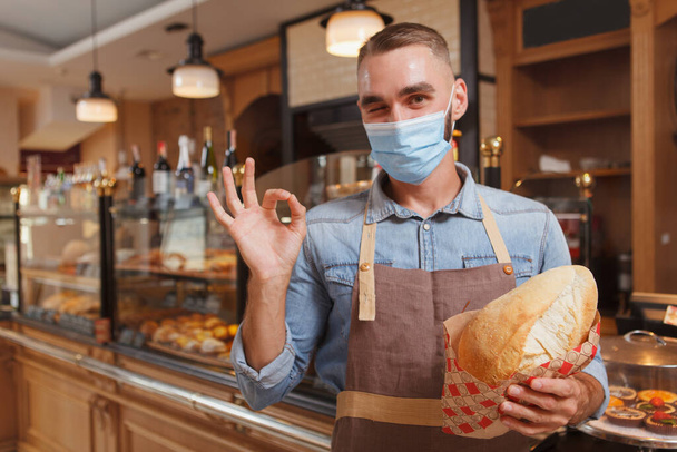 Tıbbi yüz maskesi takan erkek fırıncı, koronavirüs salgını sırasında ekmek satıyor, tamam işareti gösteriyor. - Fotoğraf, Görsel