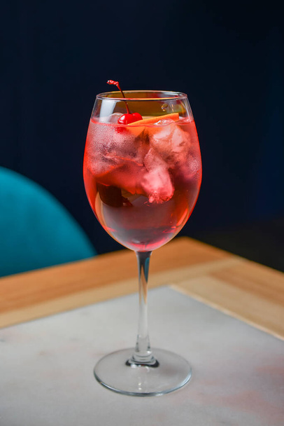 Koktajl owocowy cytrusowy w ładnym czerwonym kolorze przed czarnym tłem. Podawany w kieliszku do wina na drewnianym stole. Orzeźwiający letni napój alkoholowy z lodem i wiśnią na drewnianym stole - Zdjęcie, obraz