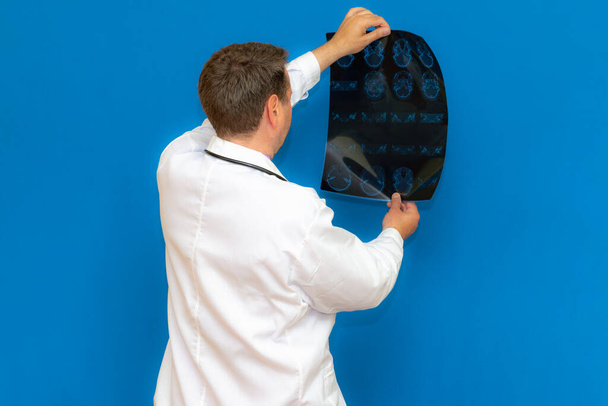 Ένας προνοητικός καυκάσιος νευρολογικός γιατρός που κοιτάζει μια αξονική εγκεφάλου σε μπλε φόντο. Braing φαντασία εξετάζεται από το νεαρό γιατρό. - Φωτογραφία, εικόνα