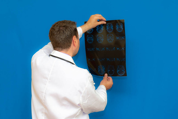 médico neurológico caucásico reflexivo mirando un escáner de ct cerebral sobre fondo azul. Braing imaginando examinado por el joven médico. - Foto, imagen