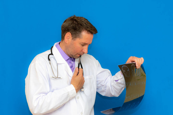 Ένας προνοητικός καυκάσιος νευρολογικός γιατρός που κοιτάζει μια αξονική εγκεφάλου σε μπλε φόντο. Braing φαντασία εξετάζεται από το νεαρό γιατρό. - Φωτογραφία, εικόνα