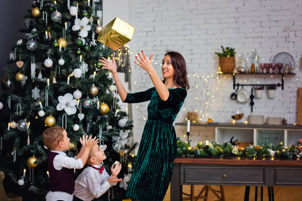 Piękna matka i dwóch chłopców obok choinki i dekoracje trzymać w rękach, wymiotować i bawić się pudełkami z prezentami - Zdjęcie, obraz