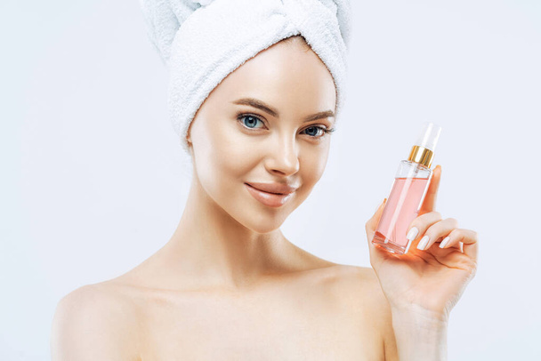 Attractive young woman applies parfum, enjoys pleasant scent, stands with naked shoulders, has natural makeup, healthy skin, wrapped towel on head after taking shower. Great aroma, try this. - Фото, изображение