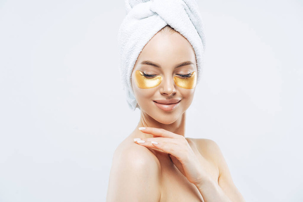 Sideways shot of tender young woman with eye collagen gold pads, healthy fresh skin, anti aging moisturising mask, touches shoulder gently, wears bath towel on head, isolated over white studio wall - Fotografie, Obrázek