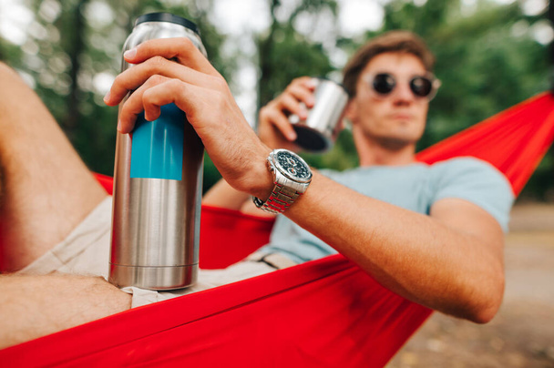 Рука молодого человека с термосом, лежащего в лесу и пьющего горячий напиток из чашки. Сосредоточьтесь на руке с серебряным термосом. - Фото, изображение