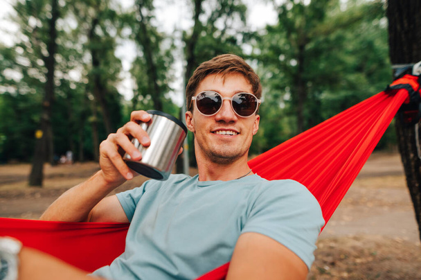 Retrato de close-up de um jovem feliz de óculos de sol deitado em uma rede com uma xícara de bebida quente na mão, olhando para o lado e sorrindo. Descanse na floresta com uma xícara de bebida quente - Foto, Imagem