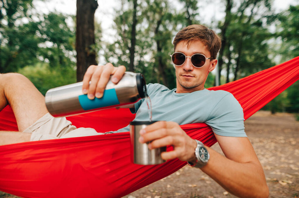 Portrait of a young man in sunglasses and light clothes lies in a red hammock and pours water from a thermos into a cup. Tourist guy uses a thermos on a hike. - Zdjęcie, obraz