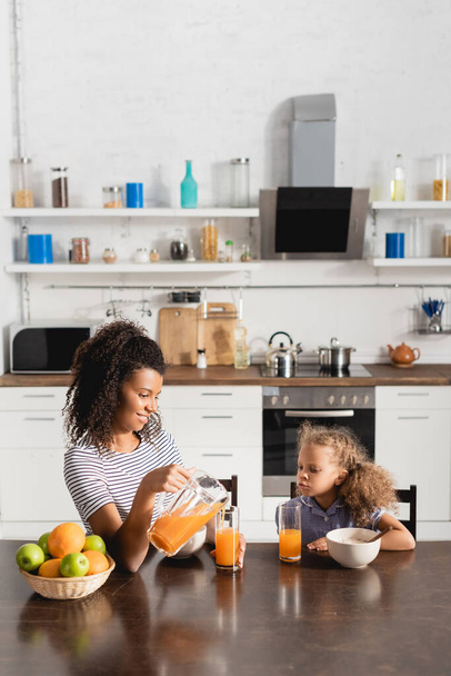 young african american woman pouring orange juice near daughter and ripe fruits - Foto, afbeelding