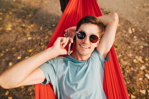 Pozitivní chlap ve slunečních brýlích leží v houpací síti na stromě v lese, mluví do telefonu a dívá se do kamery s úsměvem na tváři. - Fotografie, Obrázek