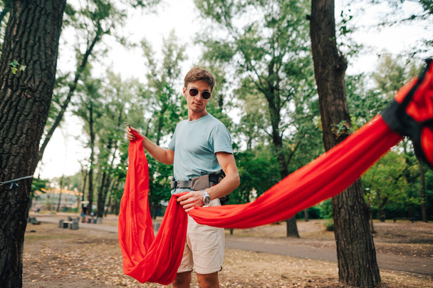 Handsome young man ties a red hammock to a tree in the park for relaxation. - Photo, Image