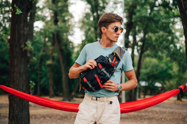 Focus op de tas. Stijlvolle jongeman in casual kleding en met een taille zak op zijn schouders staat in het bos op de achtergrond van een hangmat, kijkt naar de zijkant en poseert voor de camera. - Foto, afbeelding