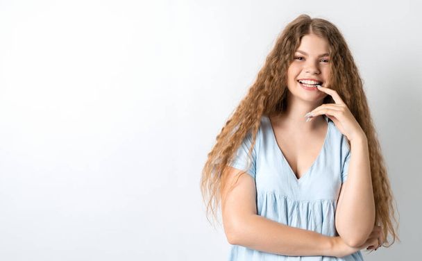Photo of thoughtful European young woman with curly long hair looks with dreamy expression aside, stands against white wall with free space for text - Zdjęcie, obraz