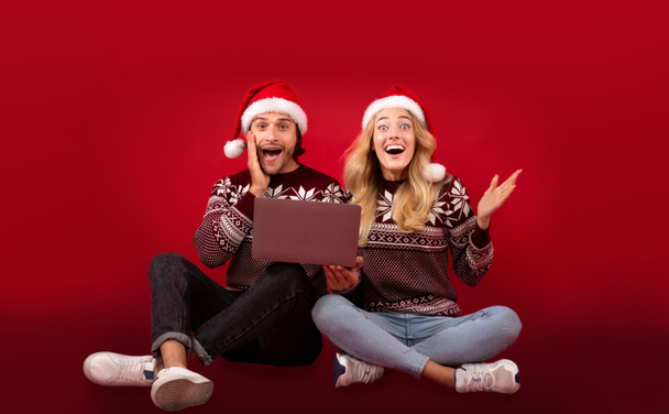Ενθουσιασμένος νεαρό ζευγάρι ψώνια για δώρα Χριστουγέννων με σούπερ εκπτώσεις στον υπολογιστή laptop πάνω από το κόκκινο φόντο - Φωτογραφία, εικόνα