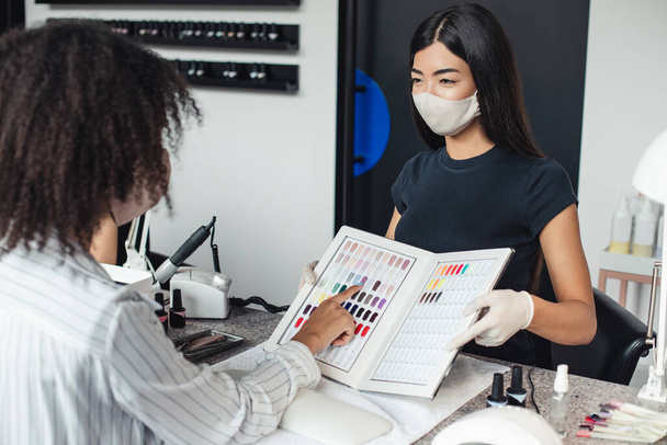 Азиатка в защитной маске смотрит на африканского американца, выбирает цвет в интерьере студии ногтей - Фото, изображение