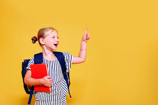 feliz niña sonriente y sorprendida en su primer día en la escuela señalando algo con su dedo, espacio amarillo para la publicidad - Foto, imagen