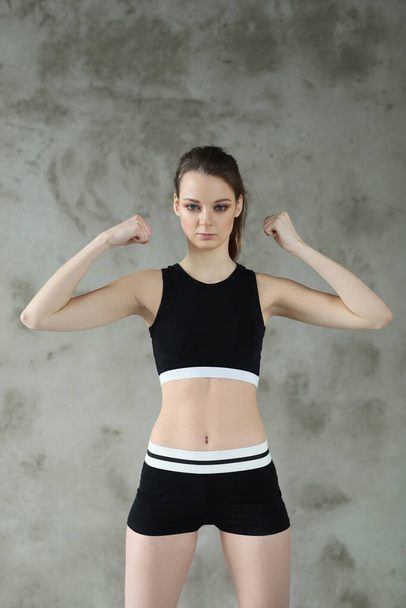 Тощая молодая женщина в спортивном костюме, позирующая на фоне серой стены, концепция спорта и тренировок  - Фото, изображение