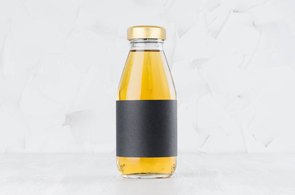 Yellow apple juice in glass bottle with gold cap and black blank label mock up on white wood table in light interior with copy space, template for packaging, advertising, design product, branding. - Foto, immagini