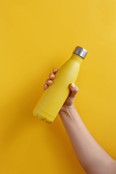 黄色の背景に黄色の再利用可能なスチールボトルを保持し、女性の手の閉じる - 写真・画像