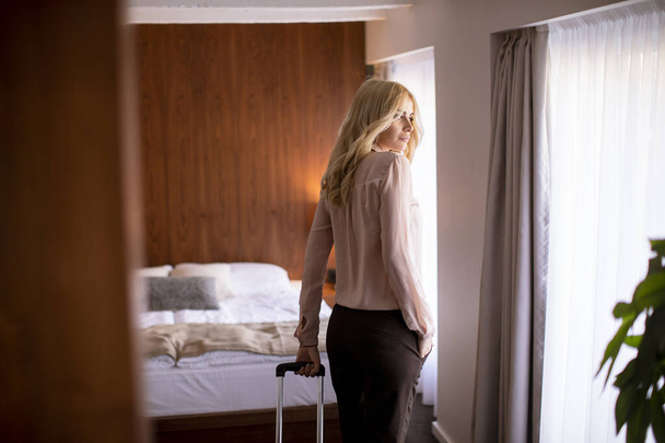 Bonita joven mujer de negocios rubia llega a una habitación de hotel con maleta roja - Foto, imagen