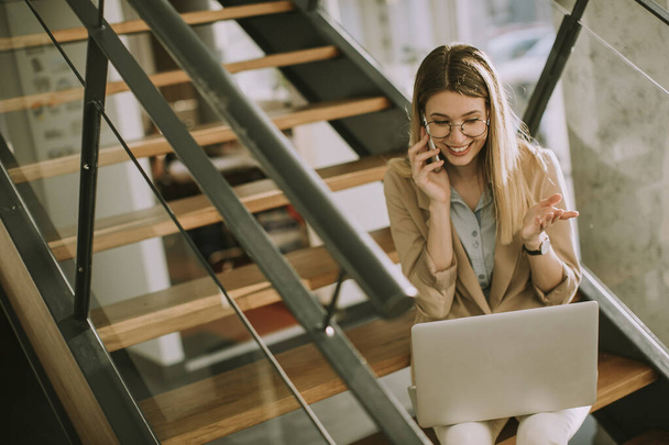 Sorridente giovane donna d'affari moderna seduta sulle scale in ufficio, che lavora sul taccuino e parla sul cellulare - Foto, immagini