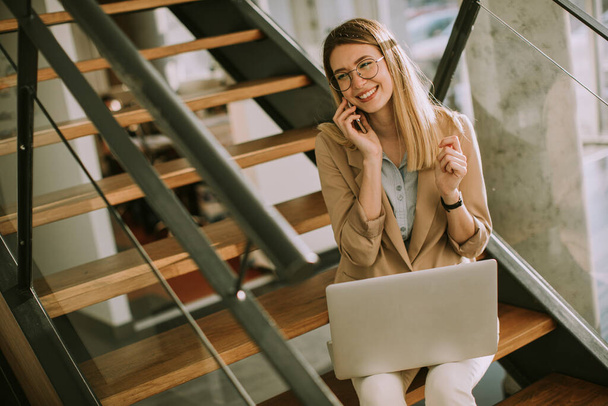 オフィスの階段に座ってノートパソコンで仕事をしたり携帯電話で話している若い現代のビジネス女性の笑顔 - 写真・画像